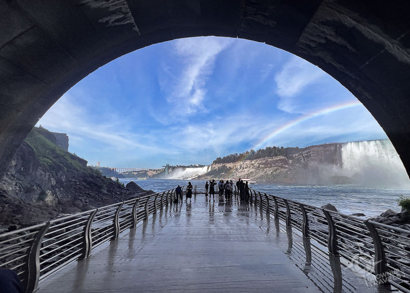 View of Niagara Falls from a pedestrian bridge in a large tunnel with a rainbow in the sky.